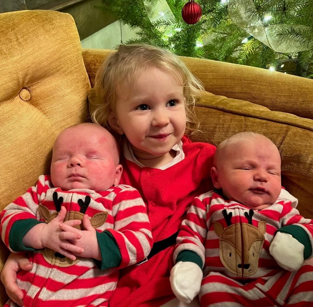 Photo from the 2022 Christmas letter: two newborn babies in matching reindeer pajamas on either side of a toddler girl on a couch by a Christmas tree