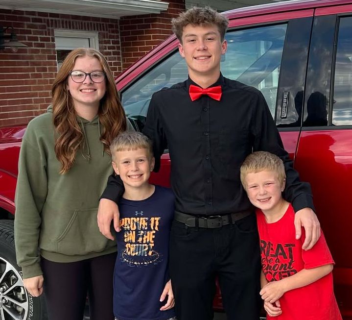 Photo from the 2024 Christmas letter: a teenage girl, a teenage boy in a red bow tie, and two younger boys standing in front of a red car